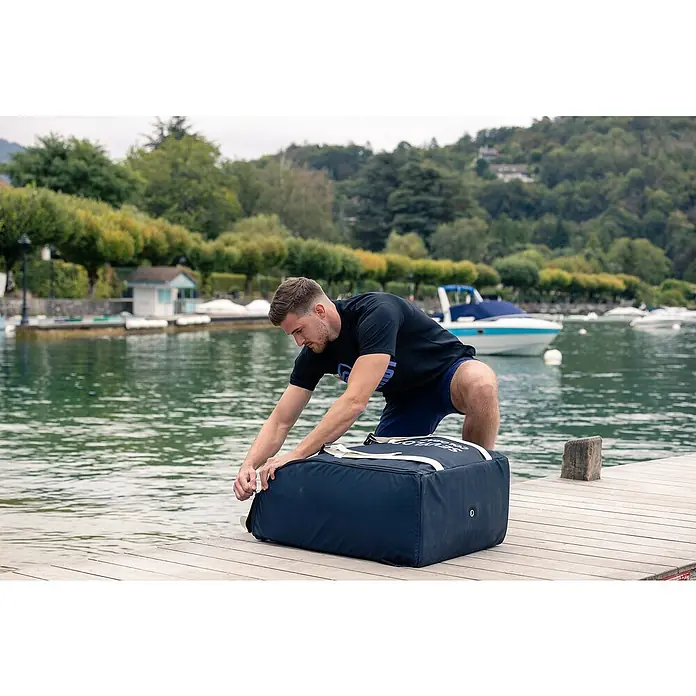 Sevylor Kajak Colorado Kit - L x B: 342 x 96 cm, Nutzlast: 180 kg, Geeignet für: 2 Personen - Ein Mann kniet auf einem Holzsteg und öffnet eine große blaue Sevylor Colorado-Tasche mit weißer Aufschrift.