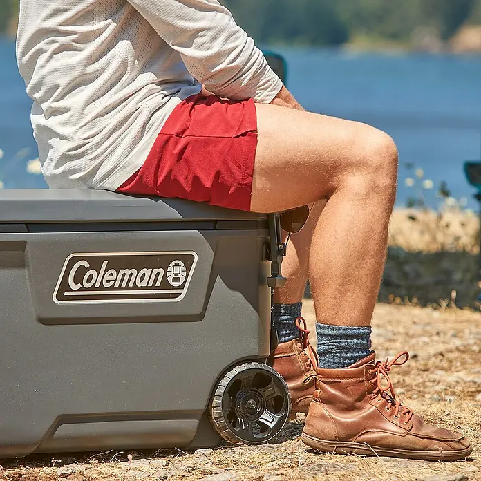Coleman Kühlbox Convoy QT65 - L x B x H: 38 x 73 x 47 cm, Schwarz, 66 l - Ein Mann sitzt auf einer grauen Coleman Kühlbox mit Rädern. Perfekt für Outdoor-Abenteuer und um Speisen und Getränke kühl zu halten.