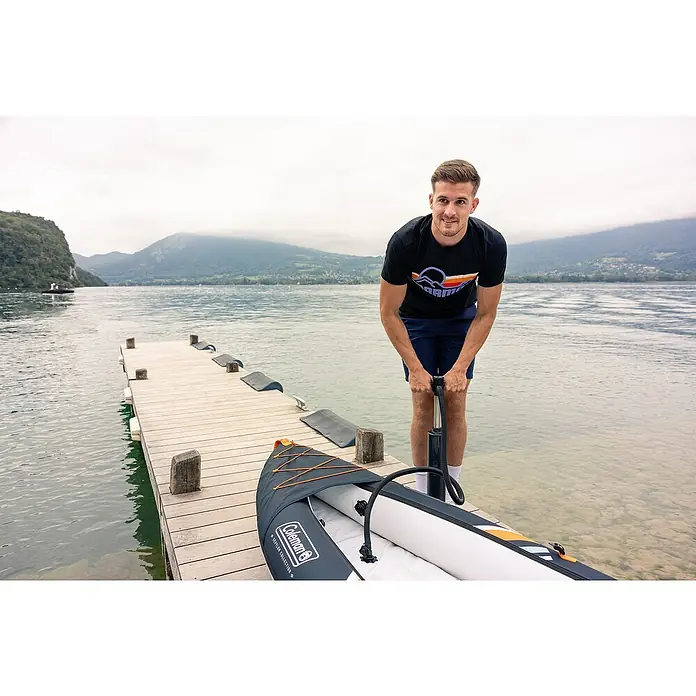 Sevylor Kajak Colorado Kit - L x B: 342 x 96 cm, Nutzlast: 180 kg, Geeignet für: 2 Personen - Mann pumpt ein Coleman Schlauchboot auf einem See auf. Das Boot ist grau und hat orangefarbene Akzente. Im Hintergrund sind Berge zu sehen.