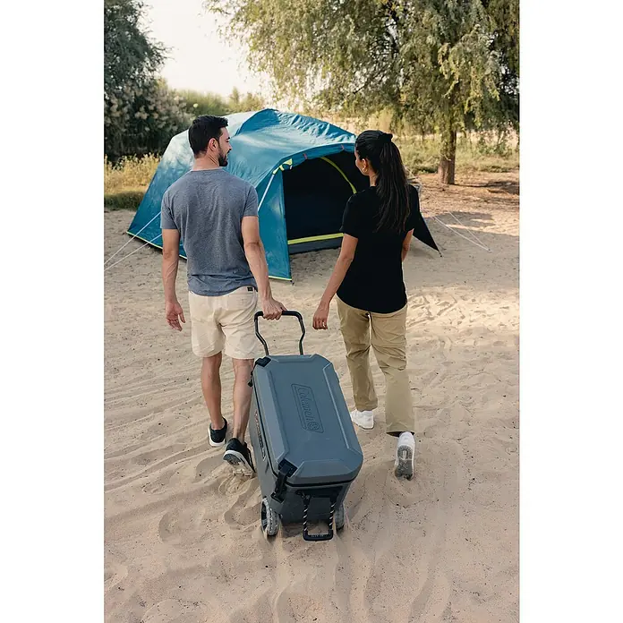 Coleman Kühlbox Convoy QT65 - L x B x H: 38 x 73 x 47 cm, Schwarz, 66 l - Ein Paar zieht einen Coleman Kühlwagen auf Rädern zum Zeltplatz. Der graue Kühlwagen ist das Hauptprodukt im Bild.