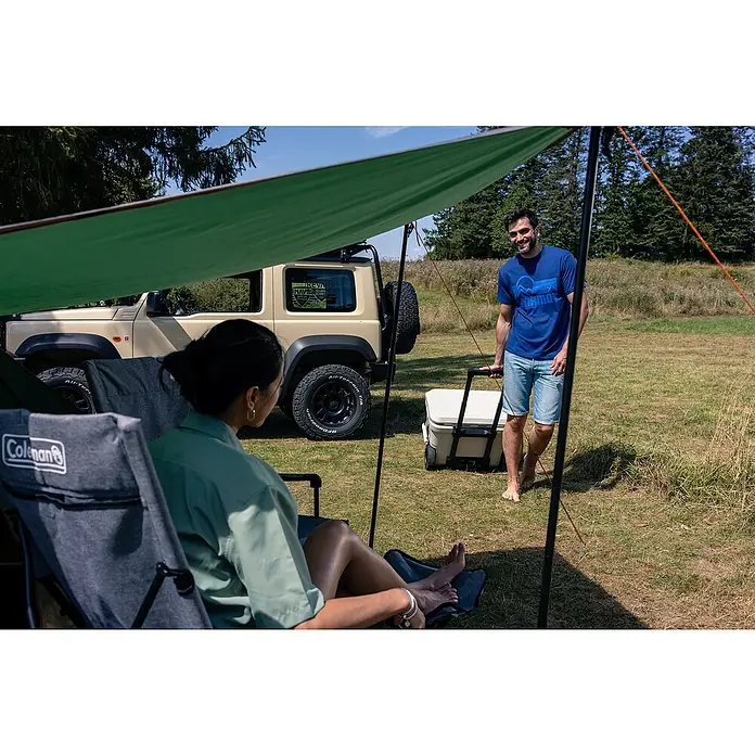 Coleman Kühlbox Pro 55QT - L x B x H: 65 x 37 x 48 cm, Schwarz, 52 l - Camping-Szene mit Coleman-Stuhl, Kühler und entspannten Campern unter einem grünen Sonnensegel.