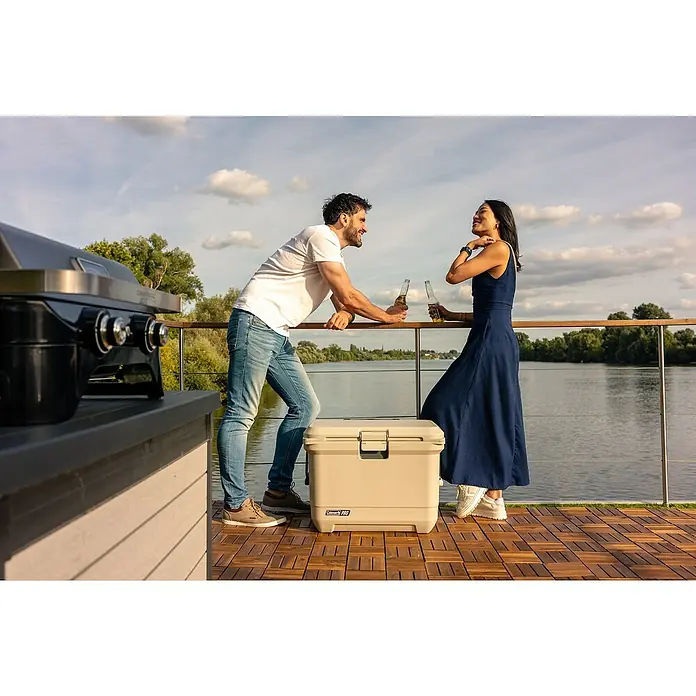 Coleman Kühlbox Pro 55QT - L x B x H: 65 x 37 x 48 cm, Schwarz, 52 l - Ein Paar entspannt sich mit Getränken neben einem Coleman Pro Kühlbox auf einer Terrasse mit Grill und Seeblick.