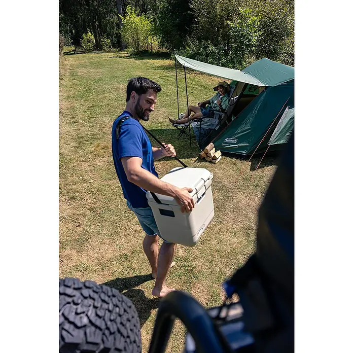 Coleman Kühlbox Pro 25QT - L x B x H: 35 x 45 x 45 cm, Weiß, 24 l - Ein Mann trägt eine Coleman Kühlbox mit Tragegurt auf einem Campingplatz, während eine andere Person sich im Hintergrund entspannt.