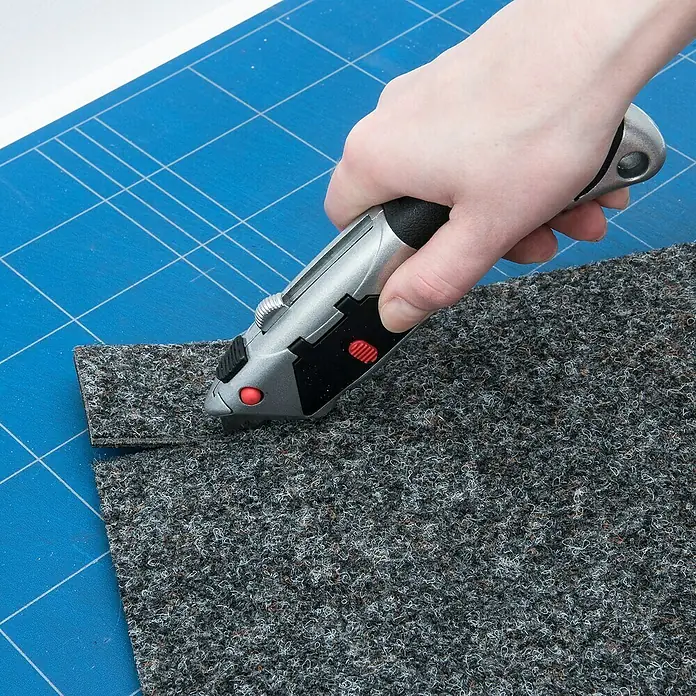 Connex Cuchillo cúter - Acero, Negro/Gris, Automático - Un cúter corta un trozo de alfombra gris sobre una alfombrilla de corte azul. El cuchillo es plateado con detalles negros y rojos.
