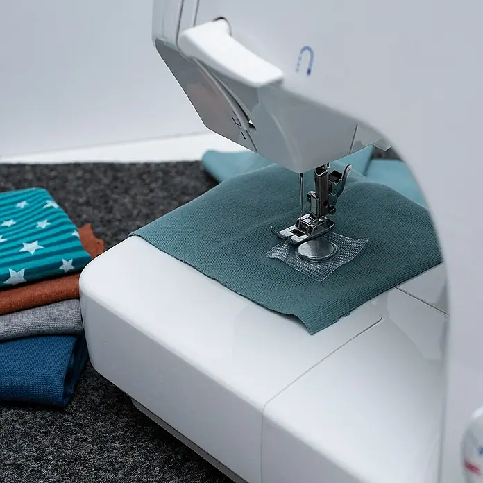 Imán - Diámetro: 18 mm, Capacidad de carga: 1,9 kg, 5 ud. - Máquina de coser blanca cosiendo tela turquesa. Al lado hay una pila de telas plegadas de colores, incluida una con estrellas.