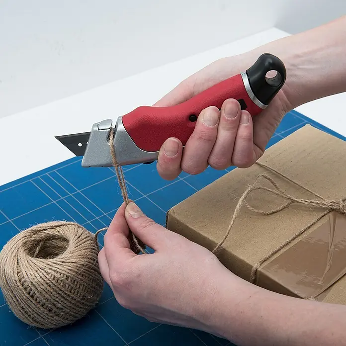 Connex Cuchillo cúter - Acero, Rojo/Negro, 3 posiciones - Imagen que muestra un cúter rojo que se utiliza para cortar una cuerda en un paquete. El cúter tiene una cuchilla negra y un mango ergonómico.
