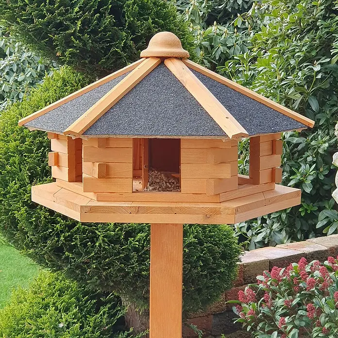 Promadino Vogelhaus Fehmarn - Holz, Hellbraun, Ausstattung: Vogelhausständer, Mit Silo - Ein detailreiches Bild eines charmanten Vogelhauses aus Holz mit einem markanten schwarzen Dach, das auf einer soliden, hölzernen Basis ruht.