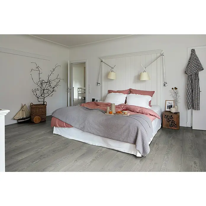 Egger Laminat NatureSense Aqua - Avery Eiche grau, Landhausdiele, 1.292 x 193 x 10 mm - Schlafzimmer mit grauem Holzboden, Bett mit grauer Decke und Frühstückstablett. Zwei Wandleuchten und ein Holzhocker als Nachttisch ergänzen das Ambiente.