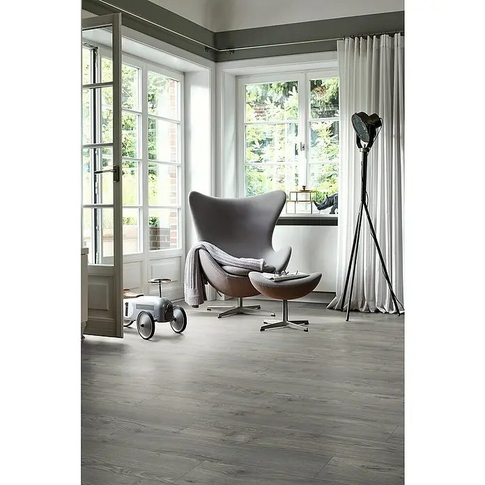 Egger Laminat NatureSense Aqua - Avery Eiche grau, Landhausdiele, 1.292 x 193 x 10 mm - Elegantes Interieur mit einem grauen Sessel, einem stilvollen Hocker und einem Vintage-Spielzeugauto auf einem modernen Holzboden.