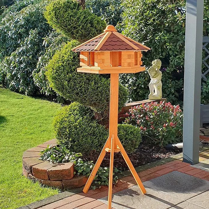 Promadino Vogelhaus Knusperhäuschen - Holz, Natur/Braun, Ausstattung: Vogelhausständer, Mit Silo - Ein Holz-Vogelhaus auf einem Ständer mit dreieckigen Beinen steht im Garten, umgeben von Grünpflanzen und Blumen.
