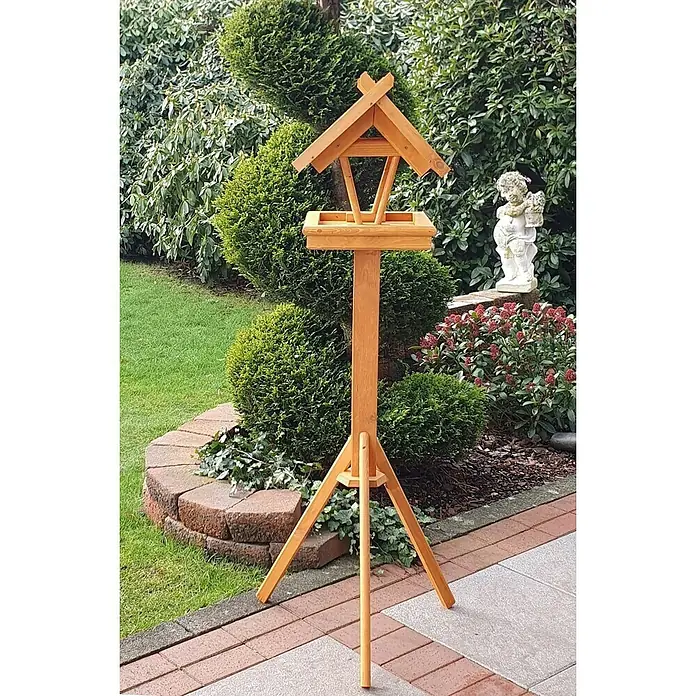 Promadino Vogelhaus Rhön - 42 x 36 x 38 cm, Holz, Braun, Ausstattung: Vogelhausständer - Eine hölzerne Vogeltränke mit einem dreibeinigen Ständer in einem Garten. Die Tränke ist von Grünpflanzen und Steinen umgeben.