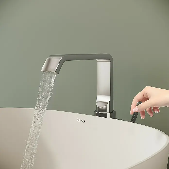 VitrA Suit Waschtischarmatur - Chrom, Glänzend, Ohne Zugstange, Höhe: 25,7 cm - Ein moderner Chrom-Wasserhahn mit fließendem Wasser in ein weißes Waschbecken; eine Hand betätigt den Griff.