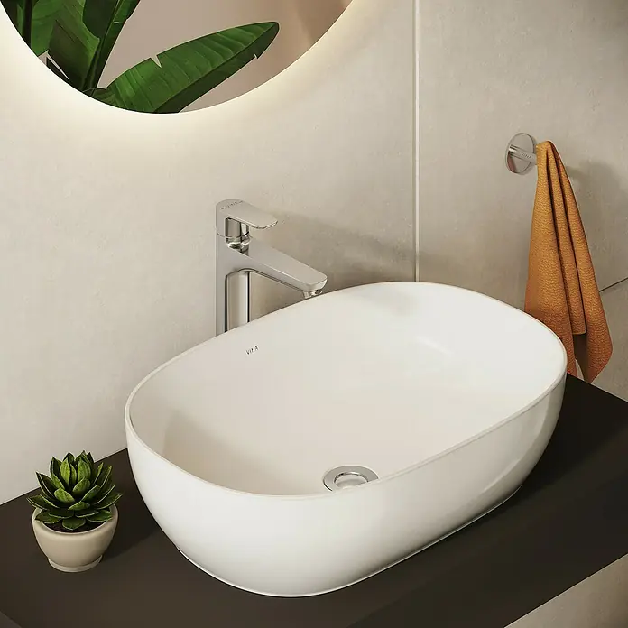 VitrA Flow Soft Waschtischarmatur - Höhe: 29,3 cm, Mit Zugstange, Chrom - Weißes ovales Keramikwaschbecken mit Chromarmatur auf dunkler Waschtischplatte in einem modernen Bad, mit Pflanze und orangefarbenem Handtuch.