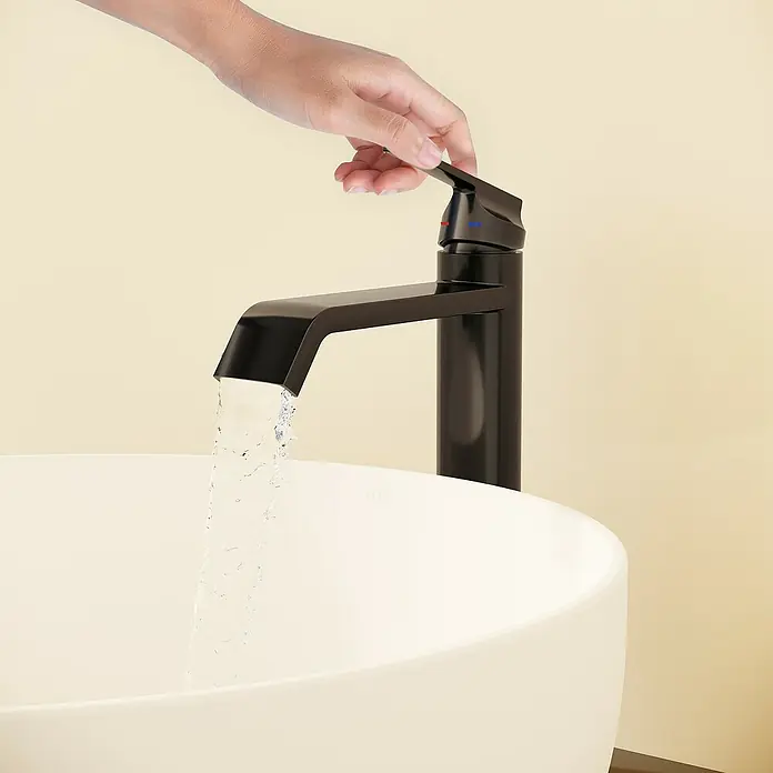 VitrA Suit Waschtischarmatur - Schwarz, Glänzend, Höhe: 26,4 cm - Hand betätigt einen mattschwarzen Wasserhahn, Wasser fließt in ein weißes Waschbecken.