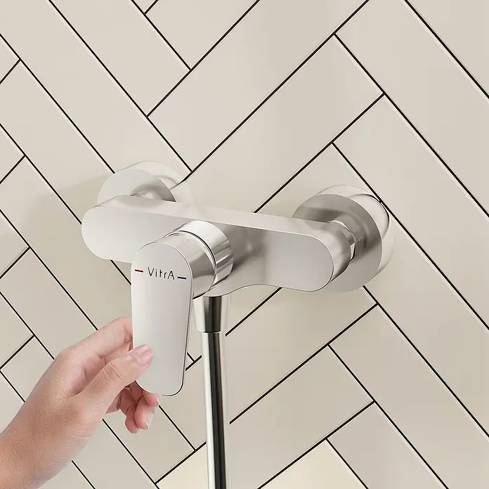 VitrA Flow Soft Brausearmatur - Ausladung: 12,7 cm, Chom, Glänzend - Eine Hand verstellt eine silberfarbene VitrA Duscharmatur an einer cremefarbenen Wand mit Fischgrätmuster.
