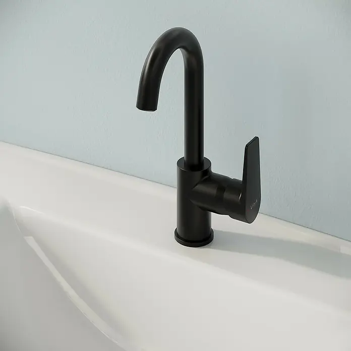VitrA Flow Square Waschtischarmatur - Schwarz, Matt, Höhe: 28 cm - Mattschwarze Einhebelarmatur mit gebogenem Auslauf, installiert in einem weißen Waschbecken vor einer hellblauen Wand.