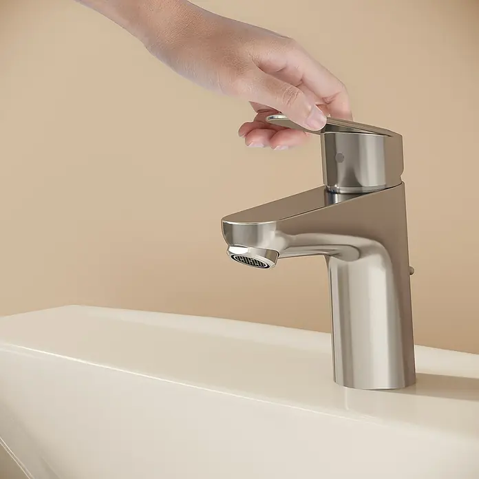 VitrA Flow Round Waschtischarmatur - Höhe: 15 cm, Ausladung Armatur: 114 mm, Chrom, Mit Zugstange - Eine Hand bedient einen Wasserhahn aus gebürstetem Edelstahl, der auf einem hellen Waschbecken installiert ist.