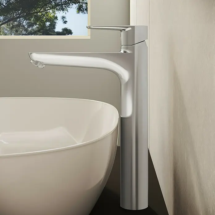 VitrA Flow Soft Waschtischarmatur - Höhe: 29,3 cm, Mit Zugstange, Chrom - Ein hoher, polierter Chrom-Wasserhahn steht neben einem weißen, modernen Waschbecken und spiegelt das Licht von einem Fenster mit grünen Bäumen wider.