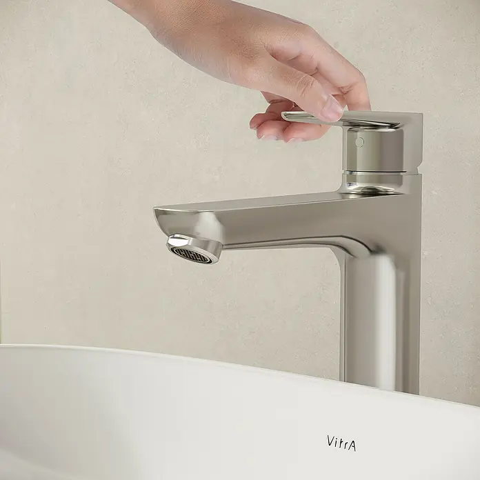 VitrA Flow Soft Waschtischarmatur - Höhe: 29,3 cm, Mit Zugstange, Chrom - Eine Hand verstellt einen eleganten silbernen Wasserhahn über einem weißen Waschbecken mit dem VitrA-Logo.