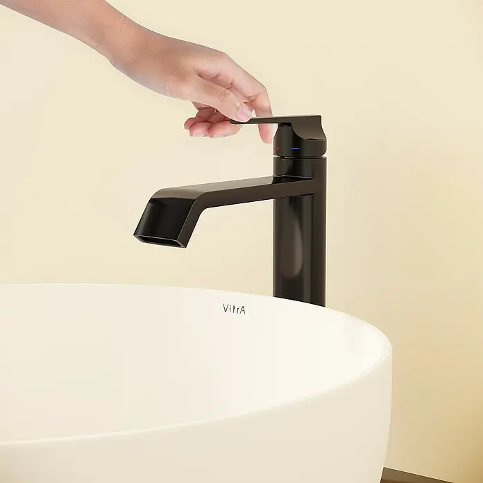 VitrA Suit Waschtischarmatur - Schwarz, Glänzend, Höhe: 26,4 cm - Hand bedient einen schwarzen Wasserhahn über einem weißen VitrA Waschbecken, Produktnutzung zeigend.