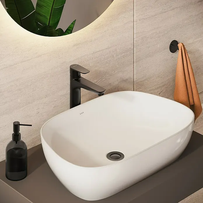 VitrA Flow Soft Waschtischarmatur - Höhe: 29,3 cm, Mit Zugstange, Schwarz - Weißes ovales Aufsatzwaschbecken mit schwarzem Wasserhahn und Seifenspender, ergänzt durch ein oranges Handtuch in einem modernen Bad.