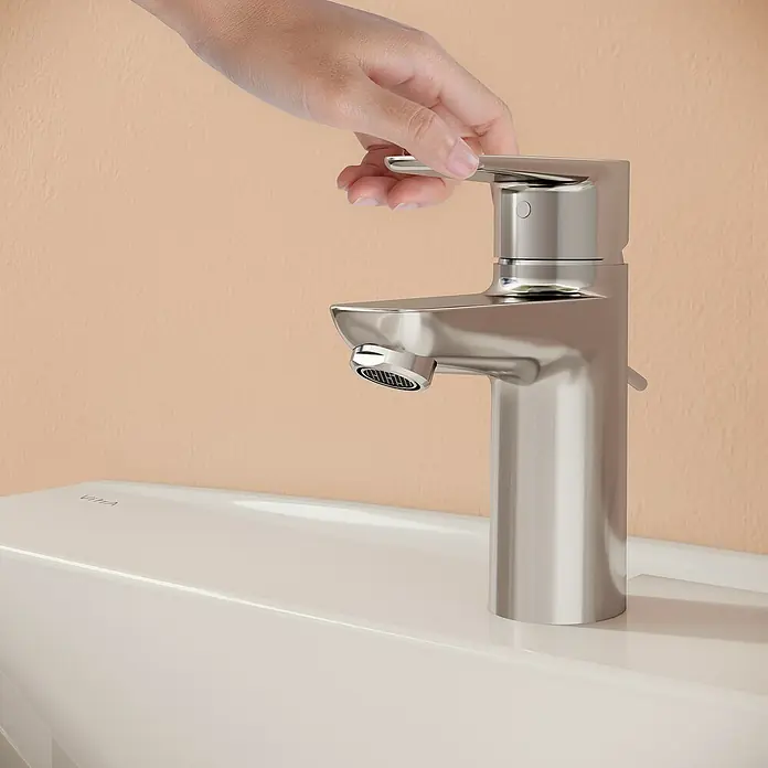 VitrA Flow Soft Waschtischarmatur - Chrom, Höhe: 15 cm, Mit Zugstange - Eine gebürstete Nickel-Einhebelarmatur auf einem weißen Waschbecken, von einer Hand bedient, vor einer pfirsichfarbenen Wand.