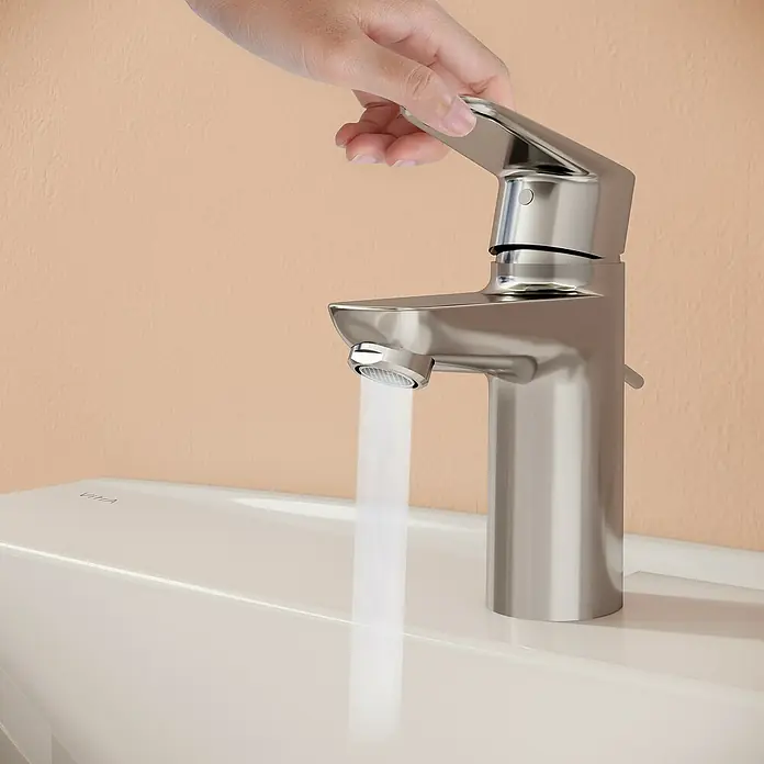 VitrA Flow Soft Waschtischarmatur - Chrom, Höhe: 15 cm, Mit Zugstange - Eine Hand dreht einen modernen silberfarbenen Wasserhahn mit fließendem Wasser über einem weißen Waschbecken.