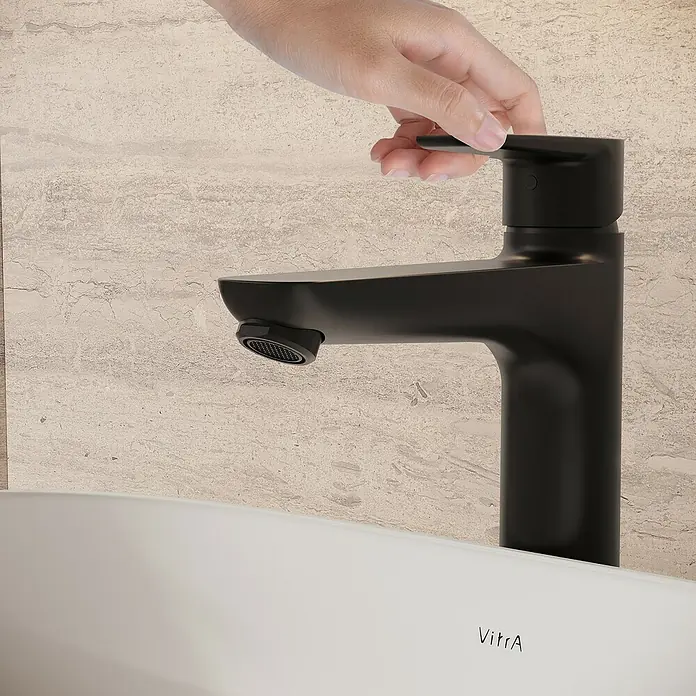 VitrA Flow Soft Waschtischarmatur - Höhe: 29,3 cm, Mit Zugstange, Schwarz - Matter schwarzer Einhebelmischer in einem Badezimmer, eine Hand verstellt den Hebel über einem weißen VitrA Waschbecken.