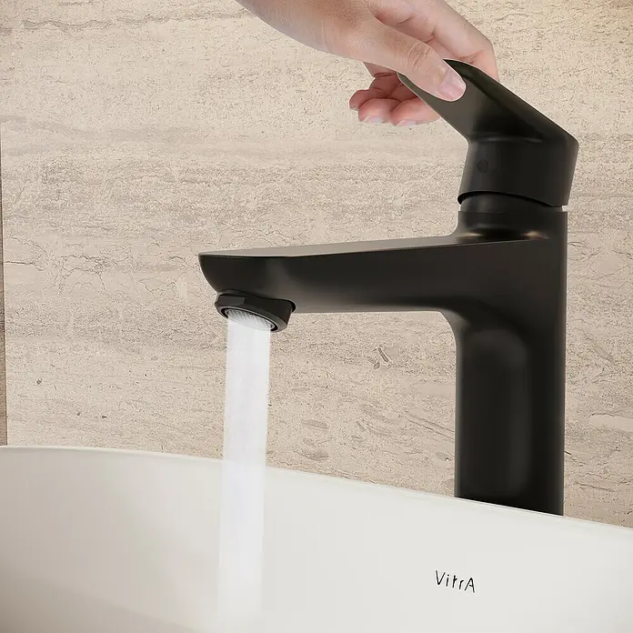 VitrA Flow Soft Waschtischarmatur - Höhe: 29,3 cm, Mit Zugstange, Schwarz - Eine Hand dreht einen mattschwarzen Wasserhahn auf, Wasser fließt in ein weißes Waschbecken vor einer hellen Steinwand.