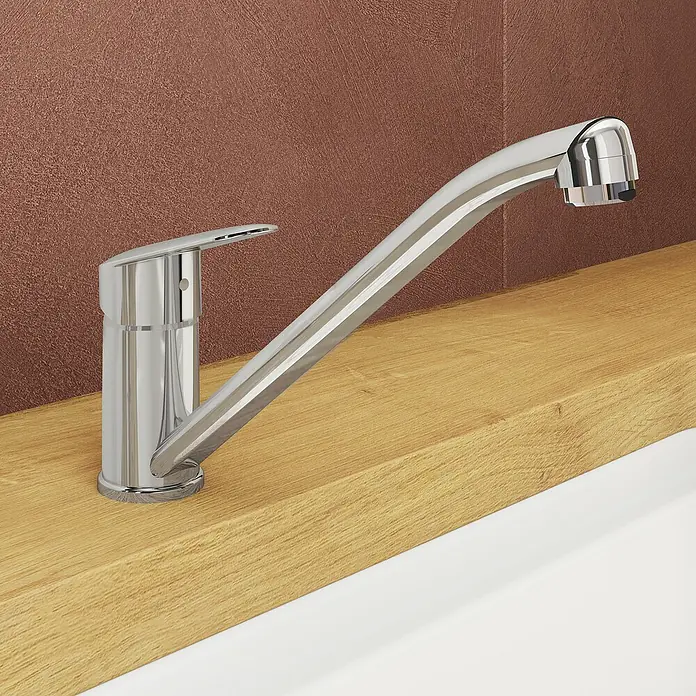 VitrA Flow Round Spültischarmatur - Höhe: 17,4 cm, Chrom, Glänzend - Verchromte Küchenarmatur mit Einhebelmischer, auf heller Holzplatte neben weißem Spülbecken vor einer strukturierten, rotbraunen Wand.