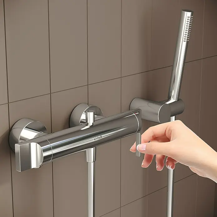 VitrA Suit Badewannenarmatur - Chrom, Glänzend - Verchromte Duscharmatur mit Handbrause. Eine Hand bedient den Mischhebel vor einer gefliesten Wand.