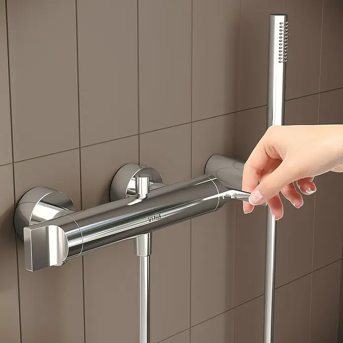 VitrA Suit Badewannenarmatur - Chrom, Glänzend - Verchromte Duscharmatur und Handbrause an einer gefliesten Wand, eine Hand regelt den Wasserfluss.