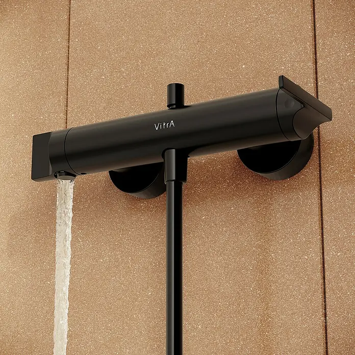 VitrA Suit Badewannenarmatur - Schwarz, Matt - Mattschwarze VitrA-Thermostat-Duscharmatur mit fließendem Wasser, montiert an einer strukturierten beigefarbenen Wand.