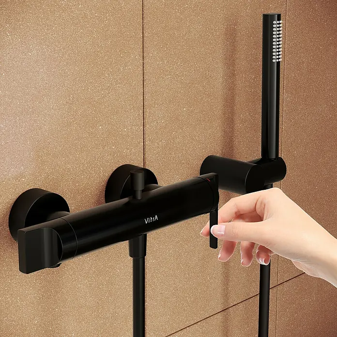 VitrA Suit Badewannenarmatur - Schwarz, Matt - Matter schwarzer VitrA Duschmischer und Handbrause an einer gesprenkelten braunen Fliesenwand, eine Hand bedient den Mischer.
