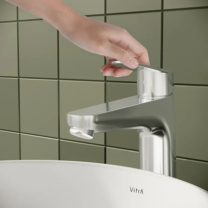 VitrA Flow Round Waschtischarmatur - Höhe: 27 cm, Ausladung Armatur: 151 mm, Chrom, Mit Zugstange - Hand dreht einen modernen Chrom-Wasserhahn über einem weißen Waschbecken auf.