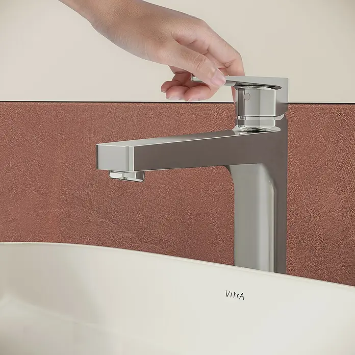 VitrA Flow Square Waschtischarmatur - Chrom, Glänzend, Höhe: 30 cm, Mit Zugstange - Eine Hand betätigt einen modernen Chrom-Wasserhahn über einem weißen VitrA Waschbecken vor einer strukturierten rotbraunen Wand.