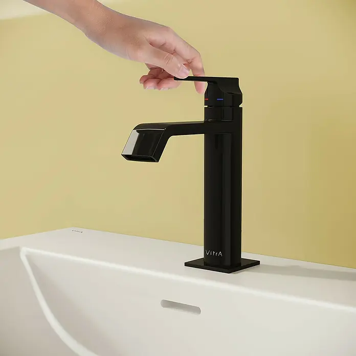 VitrA Suit Waschtischarmatur - Schwarz, Glänzend, Höhe: 19 cm - Schwarzer Wasserhahn auf einem weißen Waschbecken, eine Hand betätigt den Hebel, vor gelber Wand.