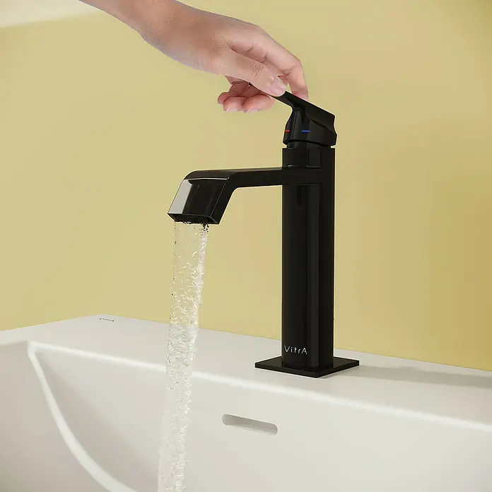 VitrA Suit Waschtischarmatur - Schwarz, Glänzend, Höhe: 19 cm - Eine Hand betätigt einen modernen schwarzen Wasserhahn an einem weißen Waschbecken, aus dem Wasser fließt.