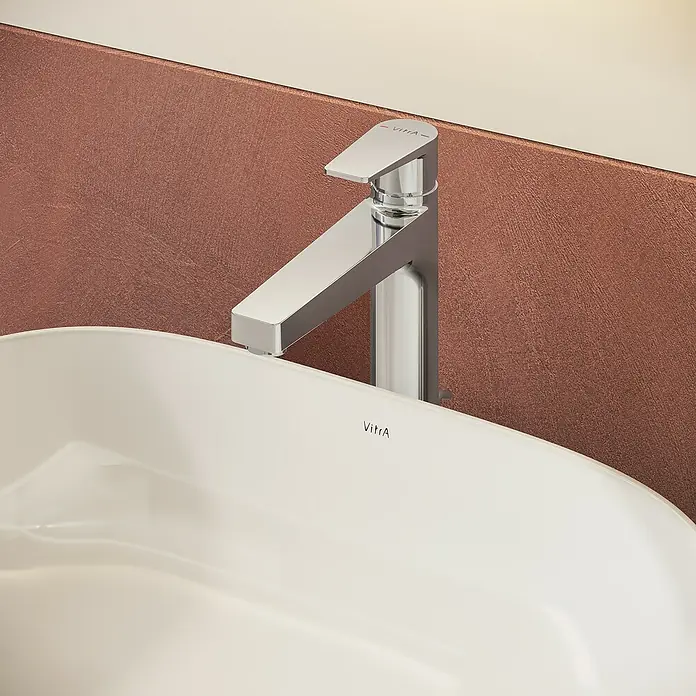 VitrA Flow Square Waschtischarmatur - Chrom, Glänzend, Höhe: 30 cm, Mit Zugstange - Moderner Chrom-Wasserhahn mit VitrA-Logo über weißem VitrA-Waschbecken, vor einer strukturierten rotbraunen Wand.