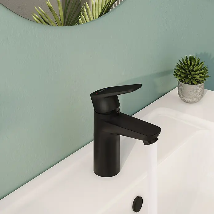 VitrA Flow Soft Waschtischarmatur - Schwarz, Höhe: 15 cm, Ohne Zugstange - Schwarze matte Armatur mit Wasserlauf in ein weißes Waschbecken vor einer türkisen Wand mit Pflanze und Spiegelreflexion.