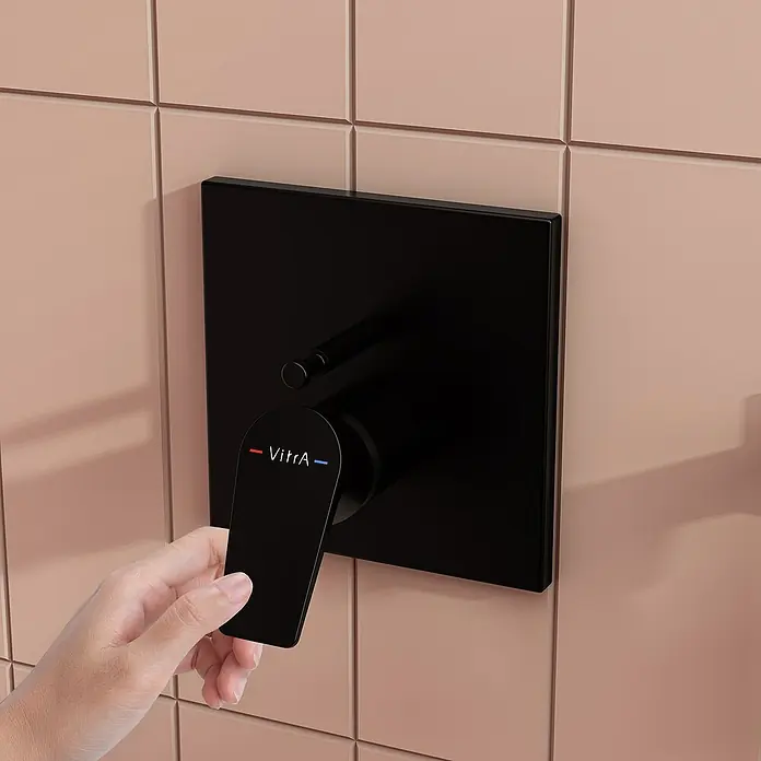 VitrA Flow Square UP-Badewannenarmatur - Ausladung Armatur: 90 mm, Schwarz, Matt - Hand betätigt einen mattschwarzen Duschmischer mit VitrA-Hebel an einer pfirsichfarbenen Fliesenwand.