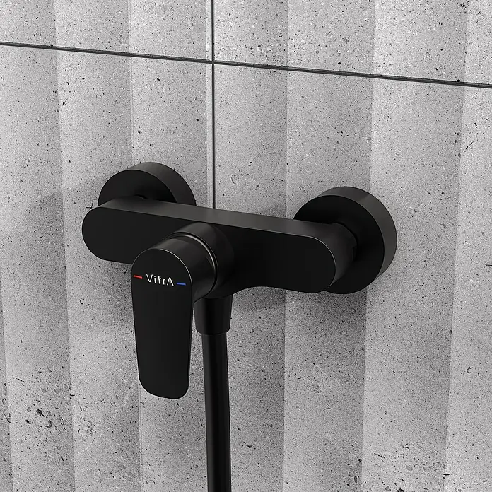 VitrA Flow Soft Brausearmatur - Ausladung: 12,7 cm, Schwarz, Matt - Mattschwarze VitrA Duscharmatur mit Griff und Schlauch, an einer grauen strukturierten Fliesenwand montiert.