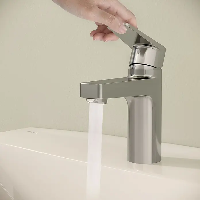 VitrA Flow Square Waschtischarmatur - Chrom, Glänzend, Höhe: 15,6 cm, Mit Zugstange - Eine Hand dreht einen modernen Chromwasserhahn auf, aus dem Wasser in ein weißes Waschbecken fließt.
