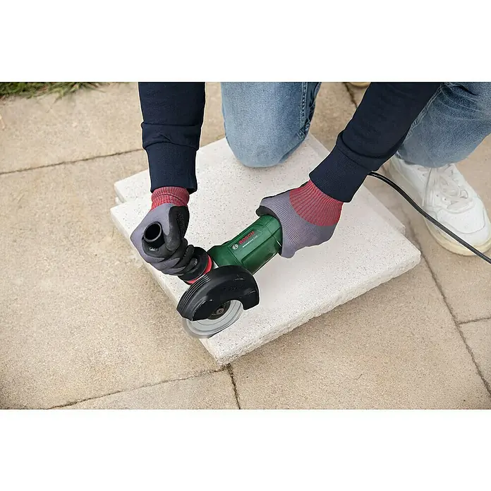 Bosch Elektro-Winkelschleifer UniversalGrind 850-125 - 850 W, Durchmesser Scheibe: 125 mm, Leerlaufdrehzahl: 12.000 U/min - Eine Person schneidet mit einer grünen Bosch-Trennschleifmaschine einen Stein auf einer Terrasse. Die Person trägt dabei Handschuhe.
