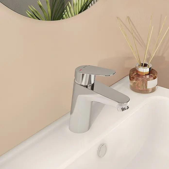 VitrA Flow Round Waschtischarmatur - Höhe: 15 cm, Ausladung Armatur: 114 mm, Chrom, Ohne Zugstange - Ein glänzender Chromwasserhahn auf einem weißen Keramikwaschbecken, mit beiger Wand, Pflanzenreflexion im Spiegel und Raumduft im Hintergrund.