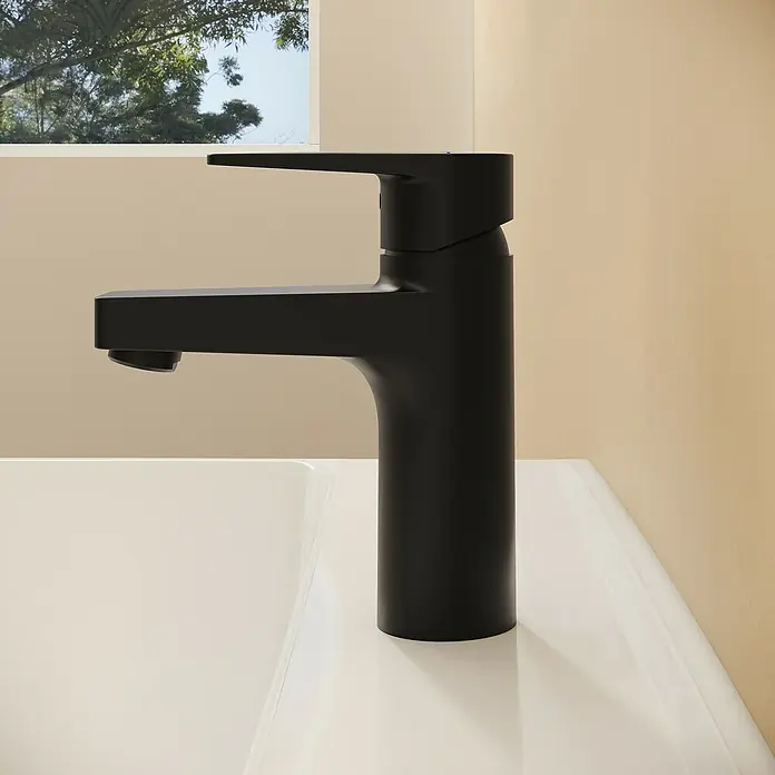 VitrA Flow Square Waschtischarmatur - Schwarz, Matt, Höhe: 15,6 cm, Mit Zugstange - Moderner, mattschwarzer Wasserhahn auf einem weißen Waschbecken in einem hellen Badezimmer mit Fensterblick auf Bäume.