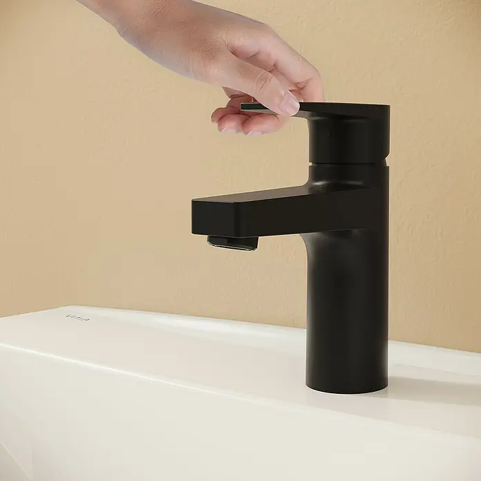 VitrA Flow Square Waschtischarmatur - Schwarz, Matt, Höhe: 15,6 cm, Mit Zugstange - Schwarze Armatur auf einem weißen Waschbecken, von einer Hand vor einer beigefarbenen Wand bedient.