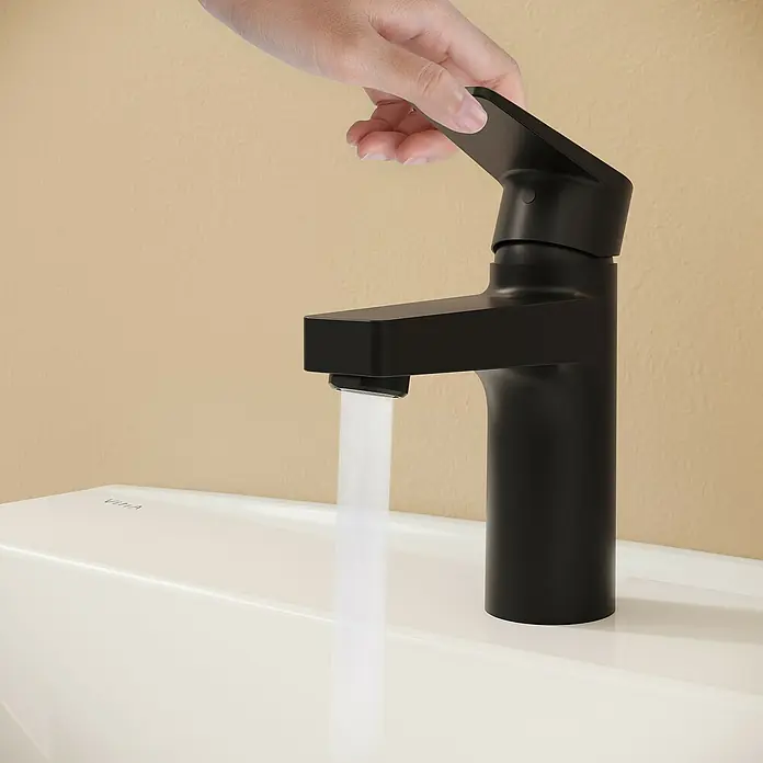 VitrA Flow Square Waschtischarmatur - Schwarz, Matt, Höhe: 15,6 cm, Mit Zugstange - Eine Hand betätigt einen mattschwarzen Wasserhahn auf einem weißen Waschbecken, aus dem Wasser fließt. Der Wasserhahn hat ein modernes Design.