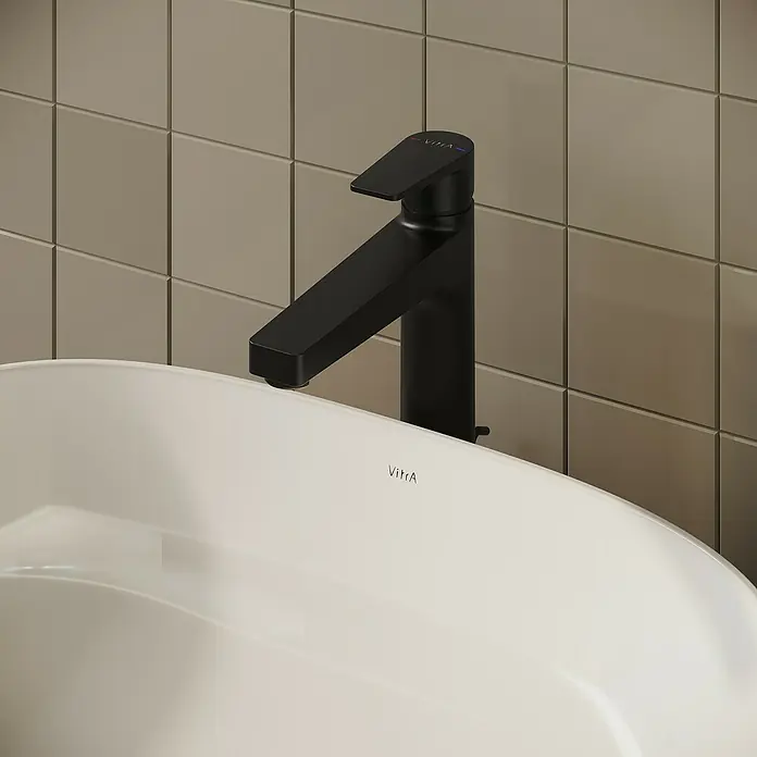 VitrA Flow Square Waschtischarmatur - Schwarz, Matt, Höhe: 30 cm, Mit Zugstange - Schwarze moderne Armatur über einem weißen Keramikwaschbecken vor beigefarbenen Fliesen.