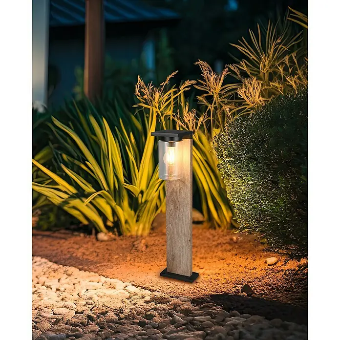 Trio Lighting Sobremuro exterior Ardila - 40 W, Color del marco: Marrón - Lámpara de jardín con textura de madera y pantalla transparente ilumina un camino de grava, rodeada de exuberante vegetación.
